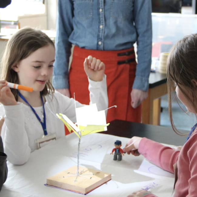 Two young children study a small model of a tree on a table