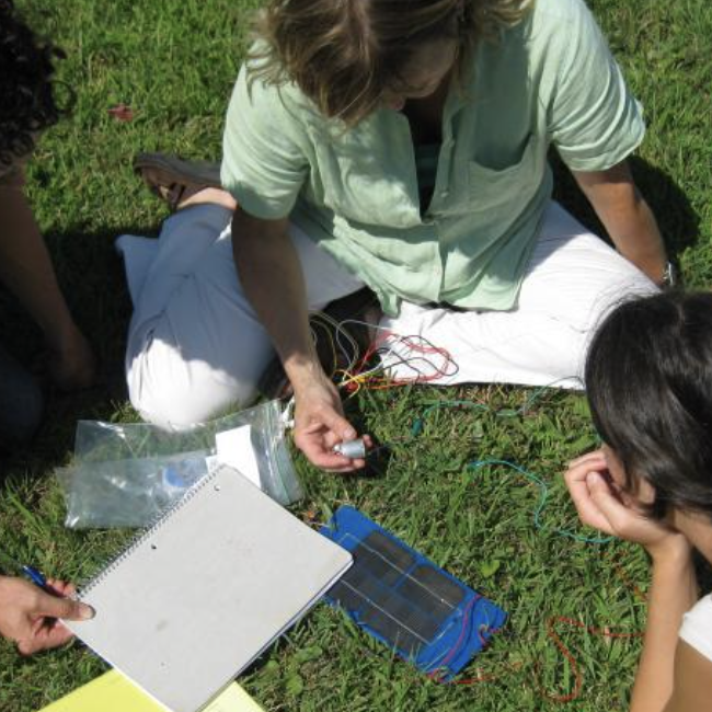 An overhead view of three people testing a small solar panel while sitting in the grass