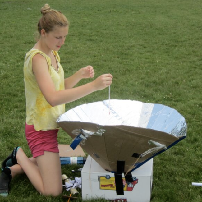 A young person holds a large metallic funnel conducting an experiment in the grass