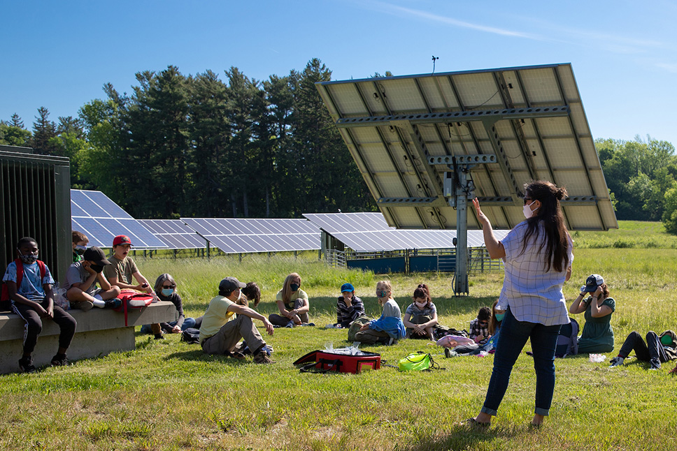 A group of students sit in the grass in a solar field as an educator gestures toward solar panels