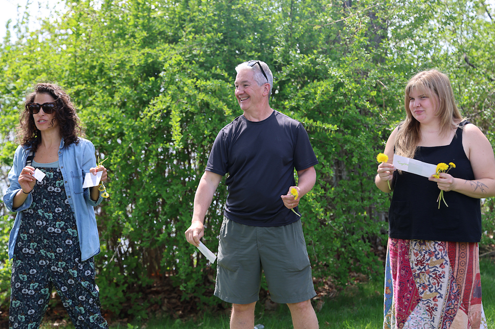 Three adults stand in a row outdoors in spring holding index cards and flowers 