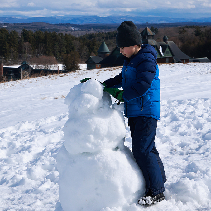 A child builds a snowman in winter with sweeping views of barn and mountains in distance