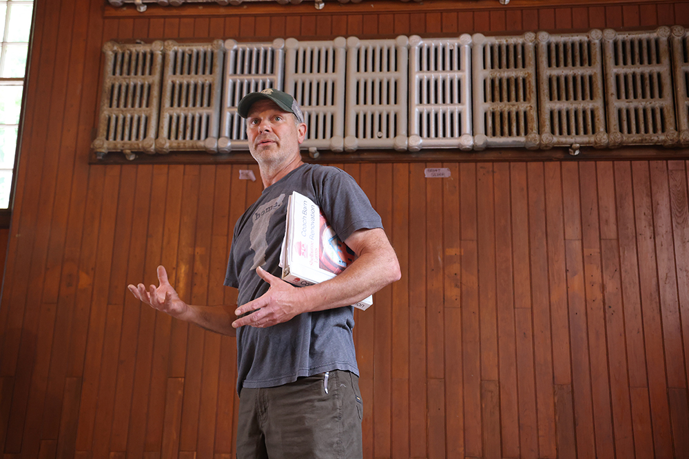 Man in gray tshirt and baseball camp with notebook under his arm gestures while talking to some people (not in frame). Behind him is a dark beadboard wall with old radiators up high