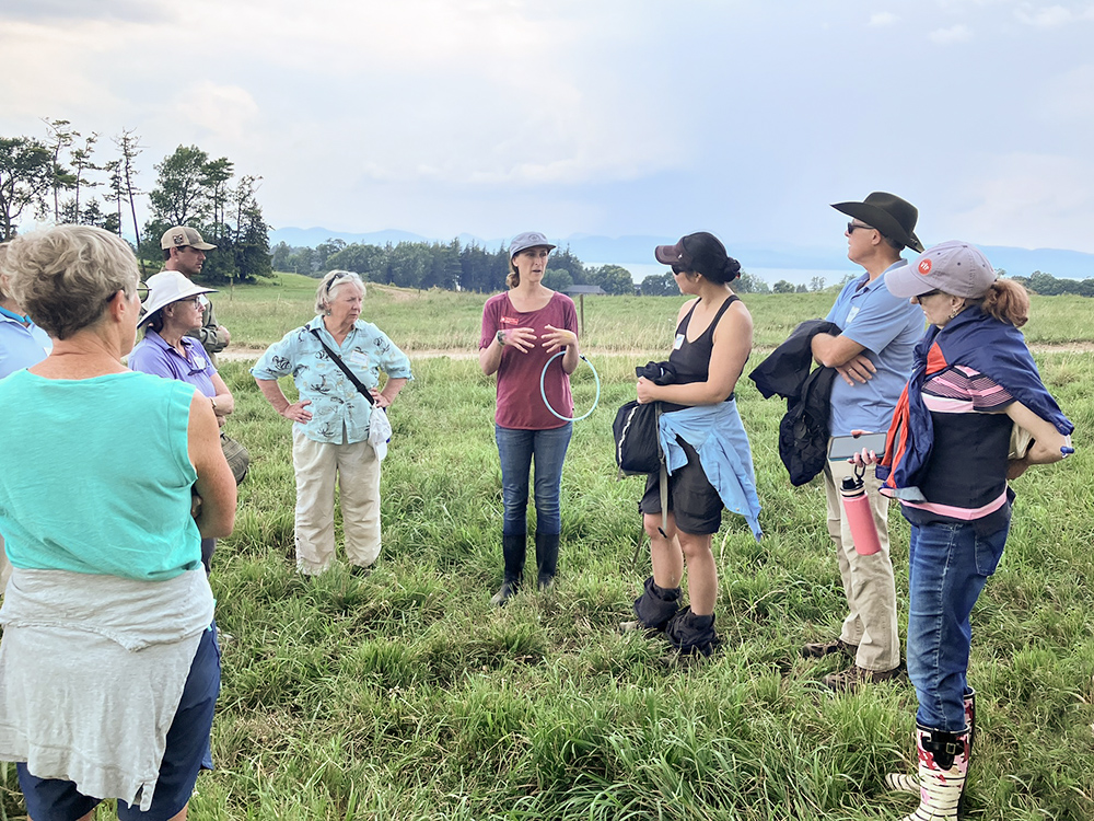 woman in jeans, farm boots and cap talking in a field to a bunch of adults.
