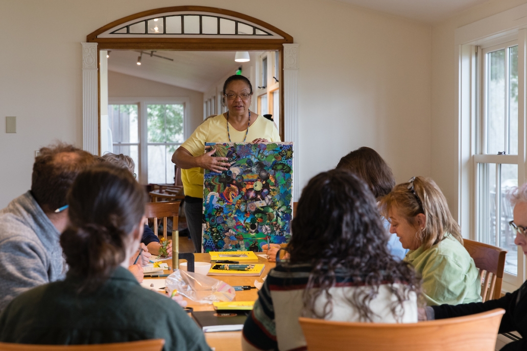A half dozen people sit around a dining room table in a cozy room with white walls, with a woman standing at the head of the table, holding a collaged piece of artwork