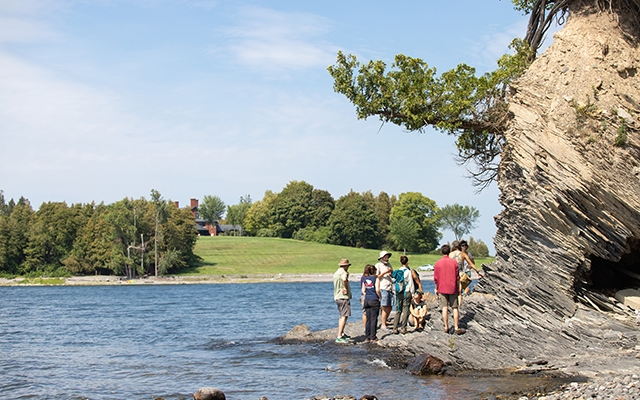 A group of a dozen people stand on the rocky shoreline of a lake under a tall cliff with rolling green hills and trees in distance