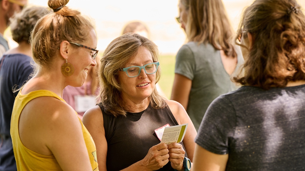 Three women stand in a circle outside in summertime looking at small flashcards