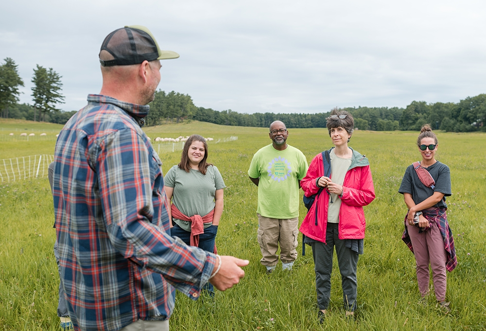 man in cap and flannel shirt facing away from camera talking to four people (long pants, short sleeves), all standing in a grass pasture.