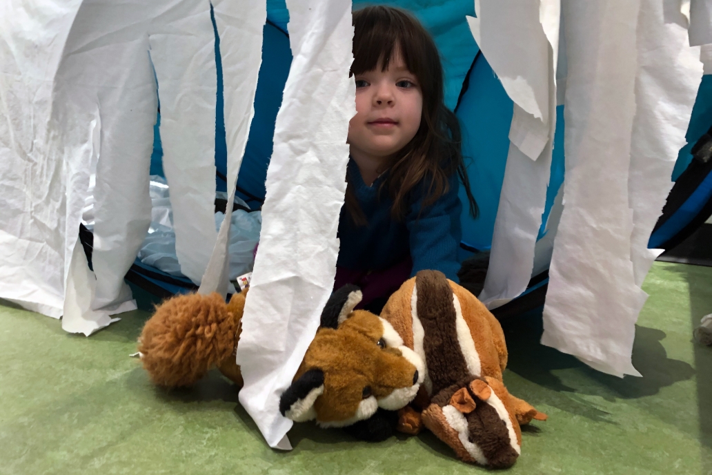 Child peeks out of a fort with hanging tissue paper sides with two fox stuffed animal toys.