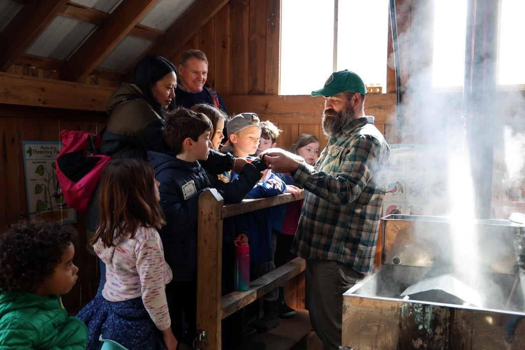 Person in sugarhouse gives a demo to a group of children and adults.