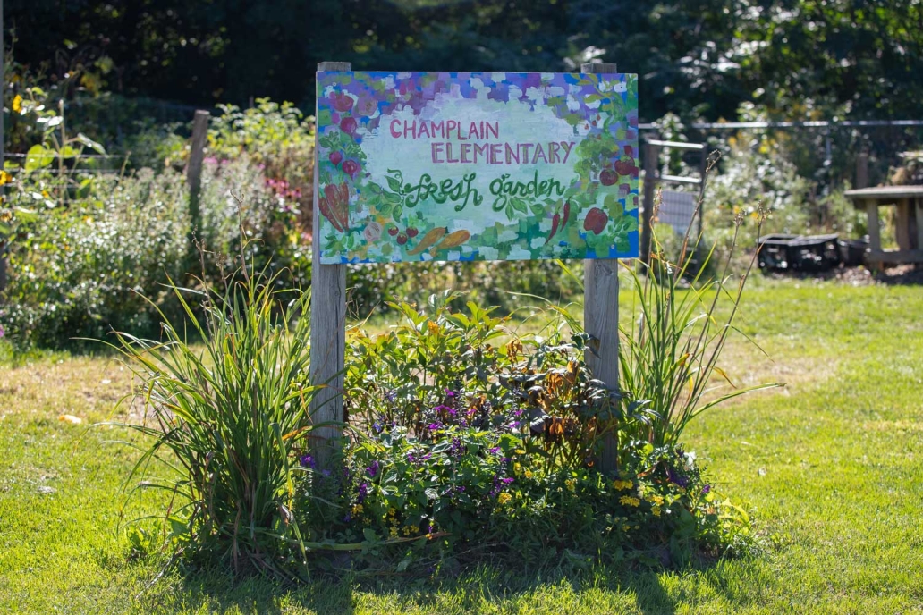 A hand painted sign with "Champlain Elementary Fresh Garden". A garden is in the background.