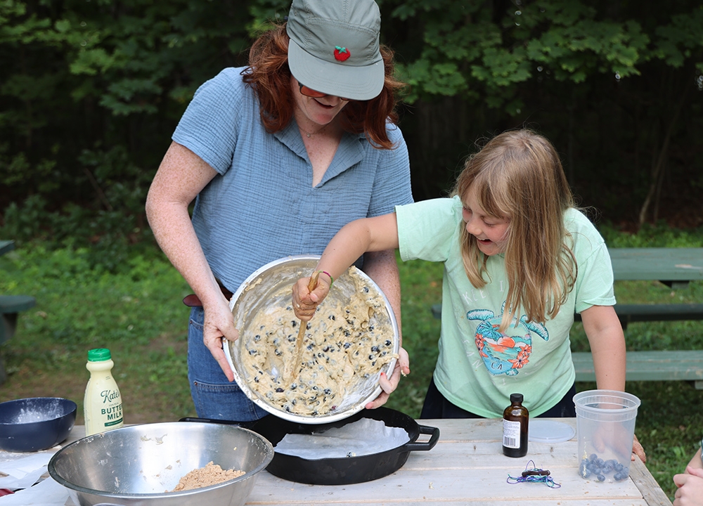 girl scrapes blueberry coffee cake dough from a bowl being held by an adult into a cast iron pan.pan