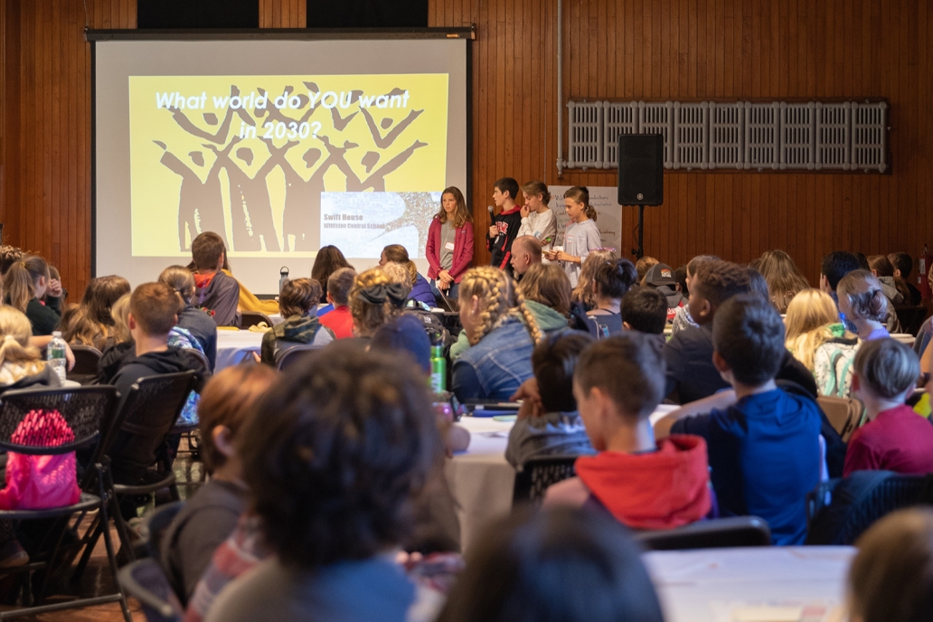 A group of young people stand in front of a projector screen that reads, what world do you want in 2030, and speak to a large room of people