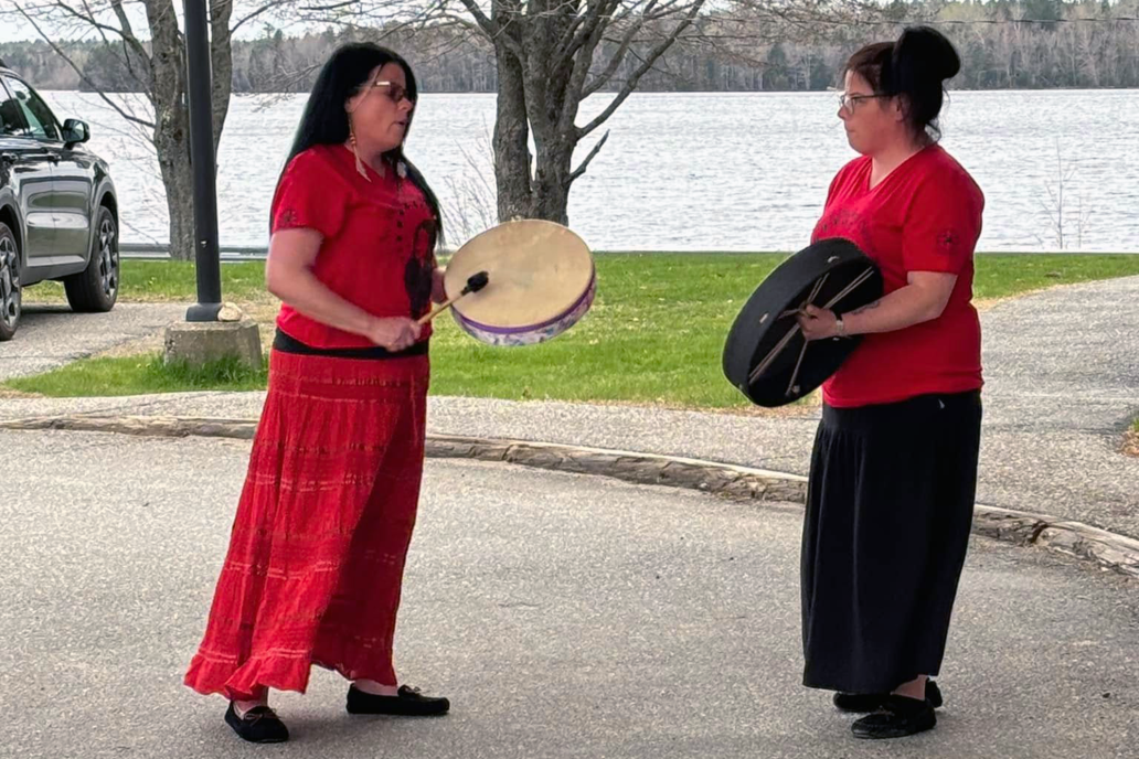 Two women wearing red playing hand drums in front of a body of water.