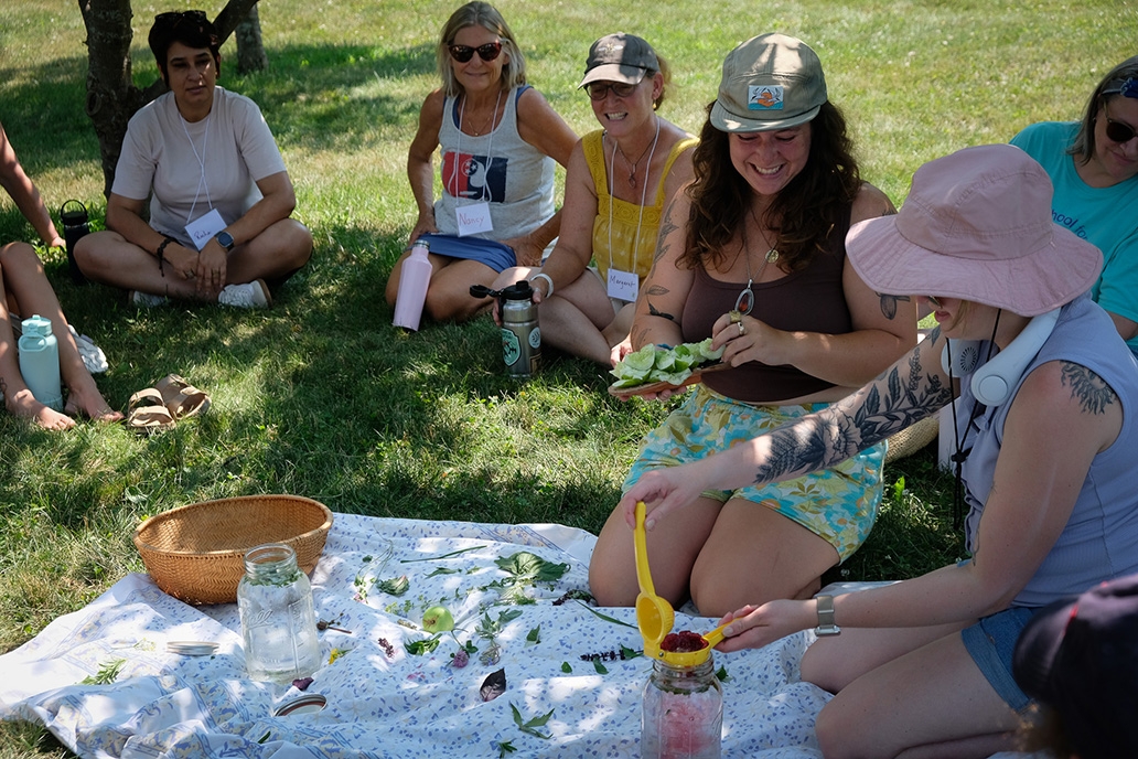 A small group of women smile while seated around a blanket in the grass, making an herbal tea during a workshop