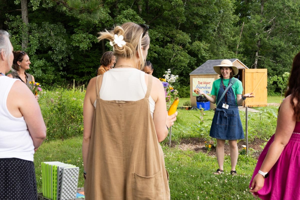 A group of people stand in a garden setting with lush greenery and a small shed in the background. One person, wearing a wide-brimmed hat and denim dress, speaks to the group.