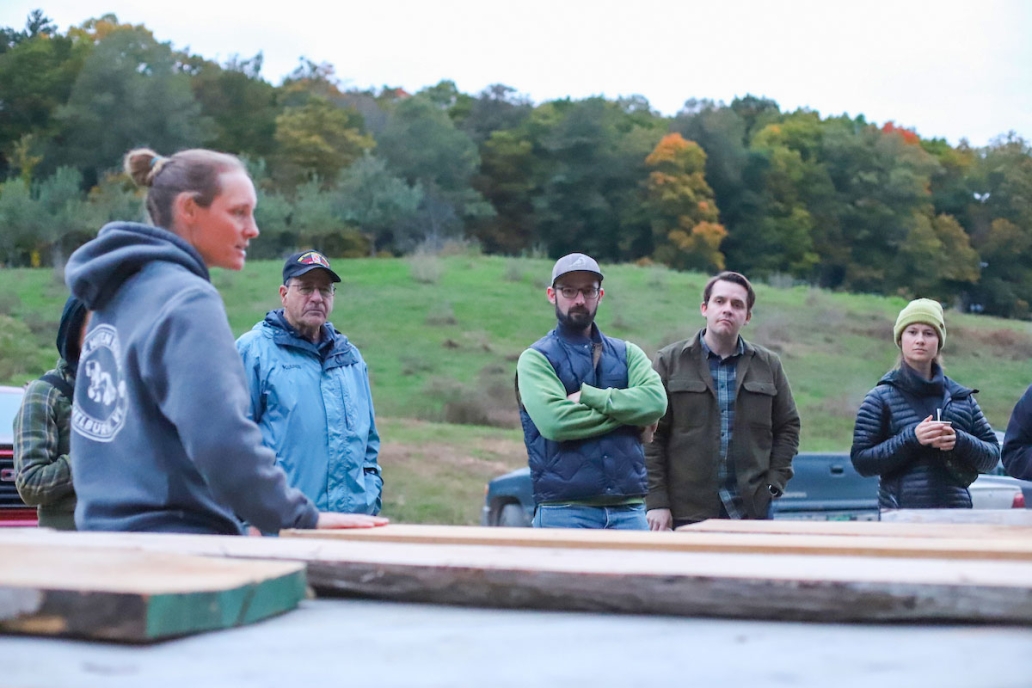 People gather around a collection of lumber outdoors, one person speaks to the group.