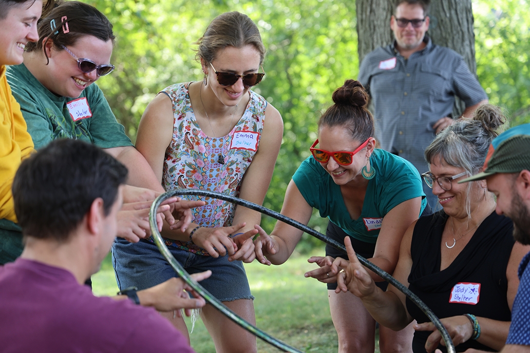 Seven people smile while balancing a hula hoop on their hands during a team building activity