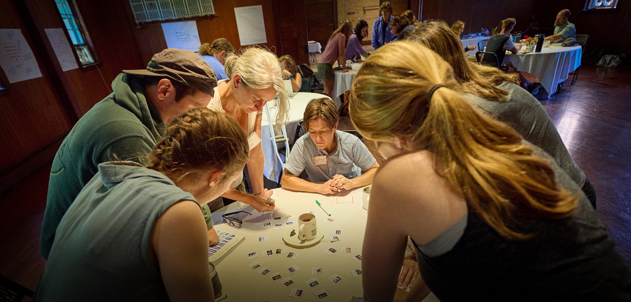 Six adults stand and sit around a circular table, looking down at small cards spread out, during a professional learning exercise