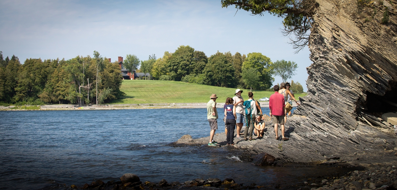 A dozen people stand on the rocky shores of a lake under a large cliff. Rolling green hills and trees stand in distance.