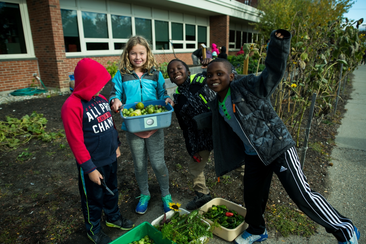 Four children excitedly participating in a garden harvest activity outside a school building.