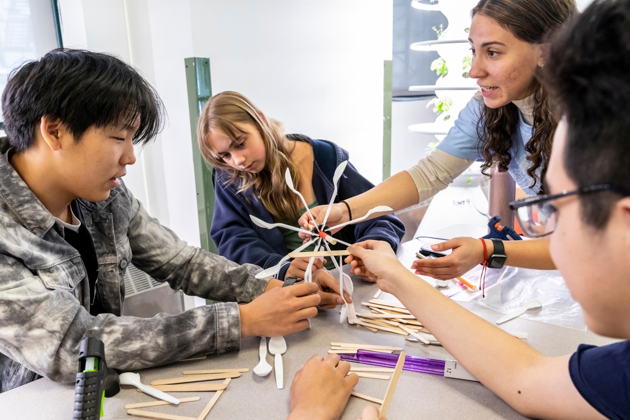 Four high school aged students sit around a table talking while working together on a miniature model of a wind turbine made of popsicle sticks and utensils