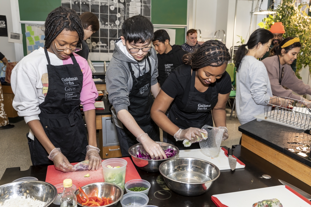 High school aged students assemble spring rolls around tables in a laboratory classroom.