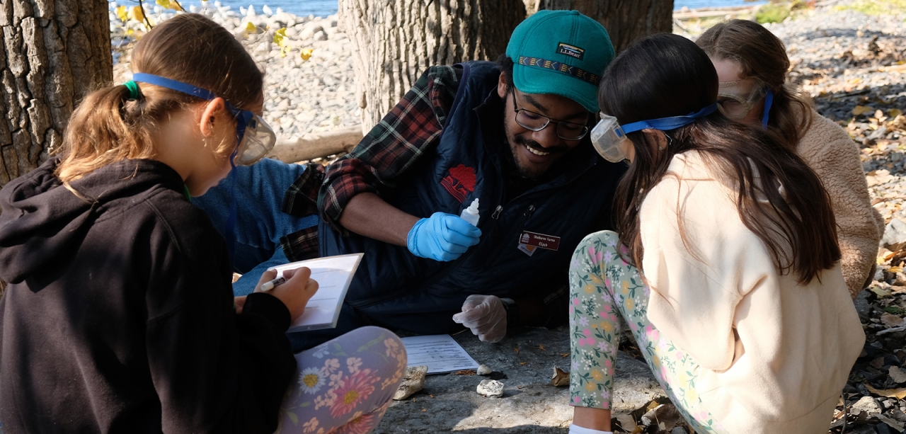 Three young students gather around an educator conducting geology experiments on a rocky lakeshore