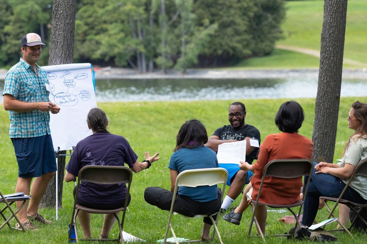 A group of educators gather outdoors, talking. One man stands next to a large easel.