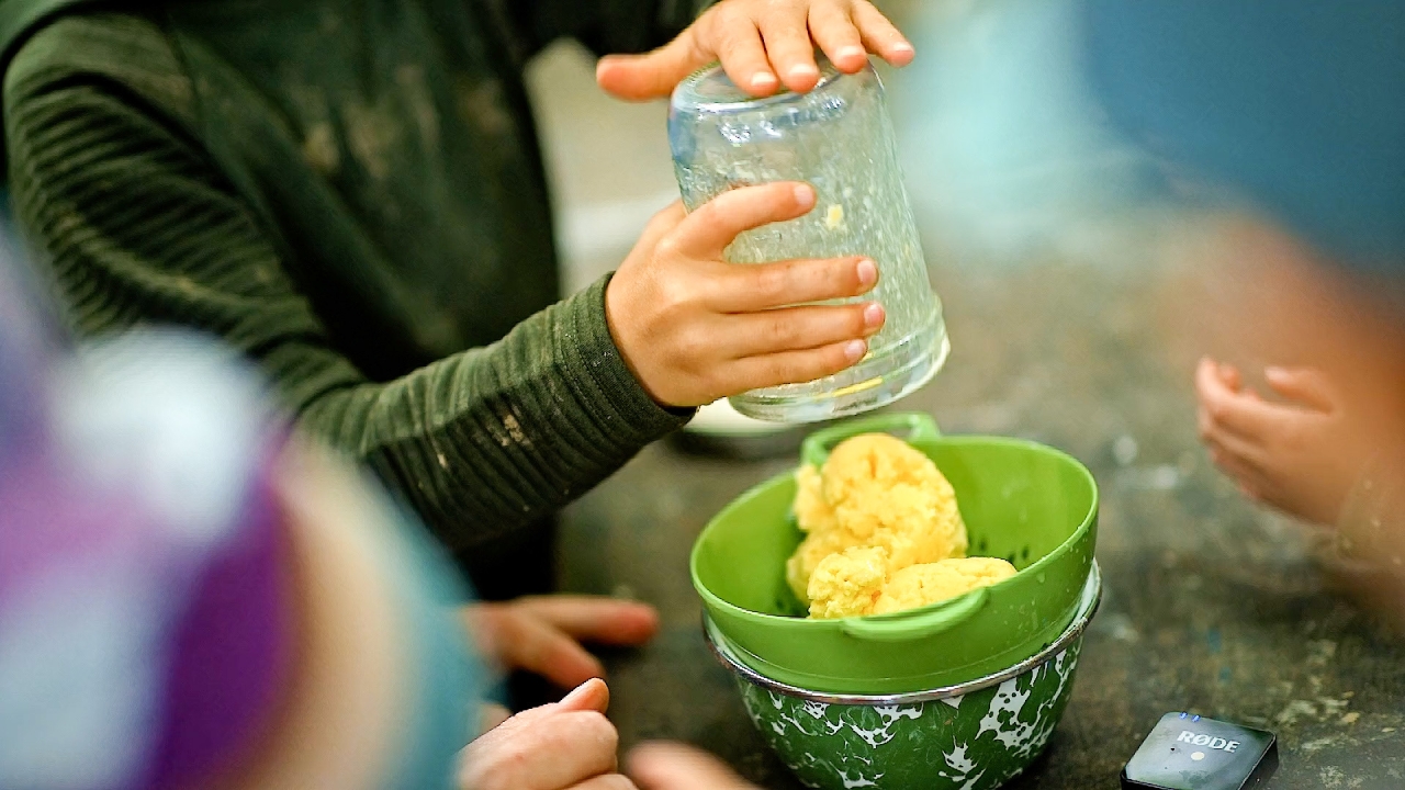 A child empties freshly made butter from a glass jar into a colander.