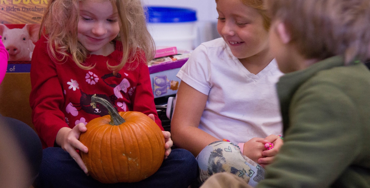 Three young students sit on a floor, excitedly examining an orange pumpkin