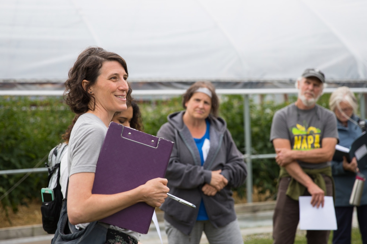 a woman with a clipboard speaks to a group of educators in a garden space