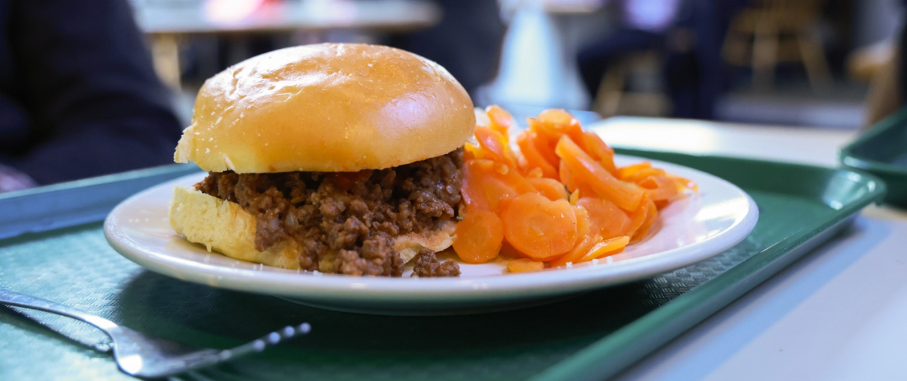 A close-up of a sloppy joe sandwich and a side of sliced carrots on a tray, set on a table in a cafeteria with people in the background.