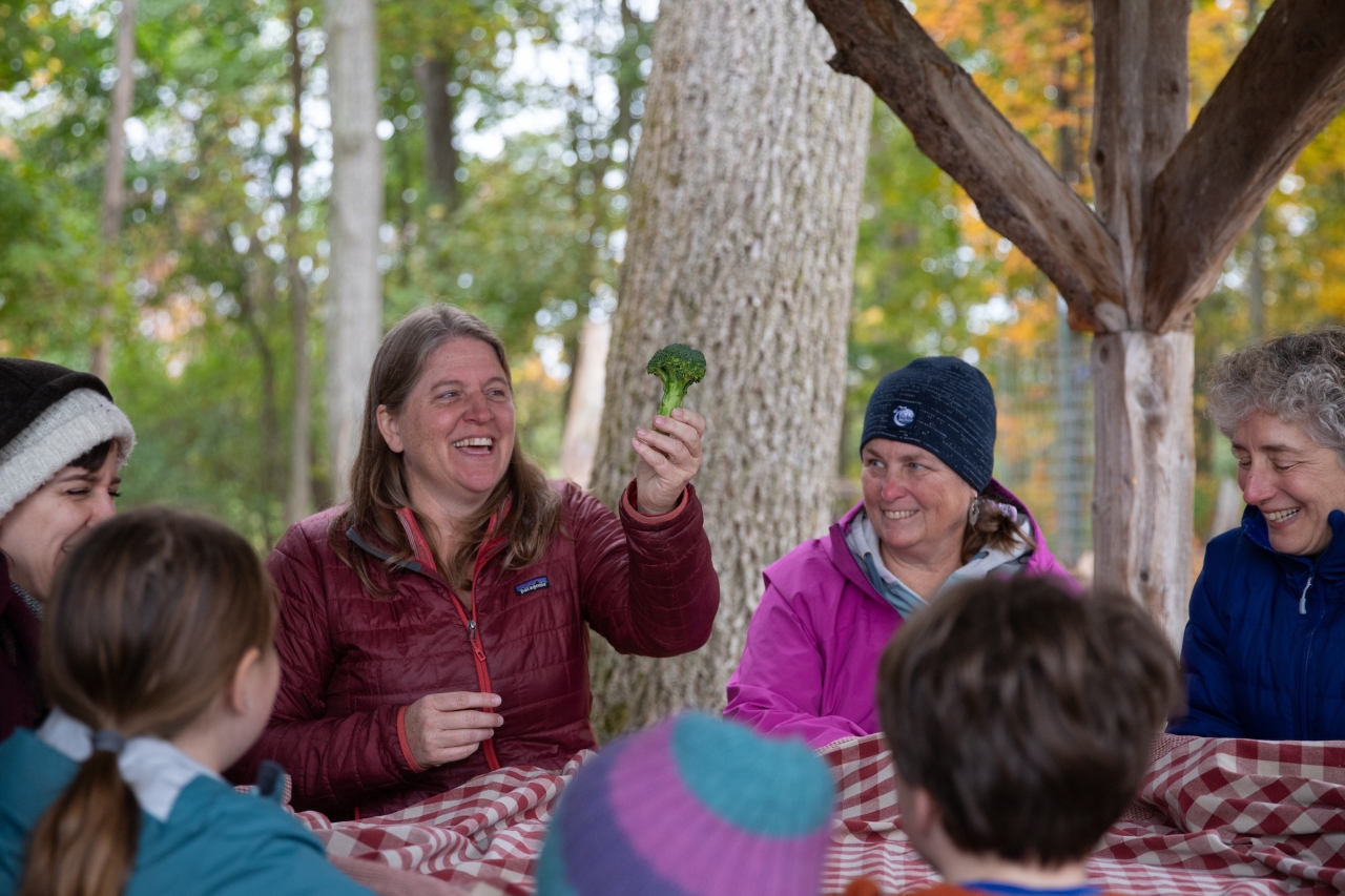 An educator holds up a piece of broccoli, other adults and children sit gathered around a picnic table