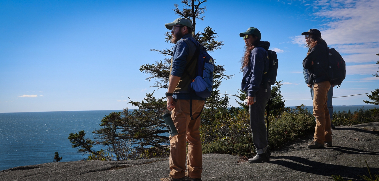 Three adults stand on a rocky mountaintop overlooking the ocean on the Maine coast