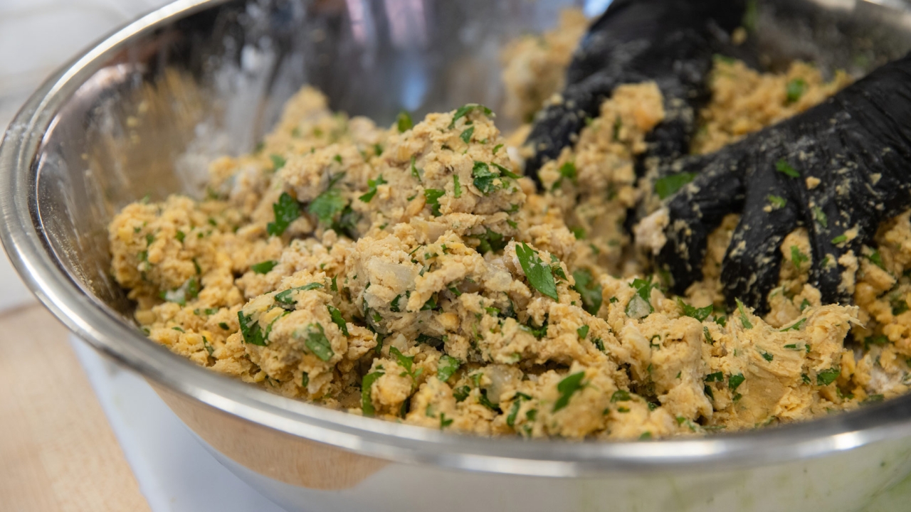 Hands wearing black gloves are mixing a large bowl of seasoned chickpea mixture with herbs.