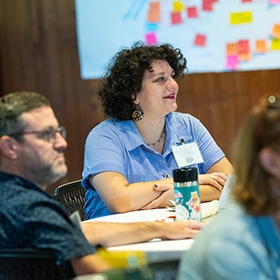 Three educators sit in the Shelburne Farms Coach Barn watching a presentation. Educator at center smiles. Post-it notes are stuck on the wall in background.