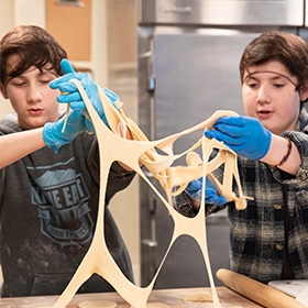 Two teenage children wearing gloves and hairnets stretch dough in an industrial kitchen