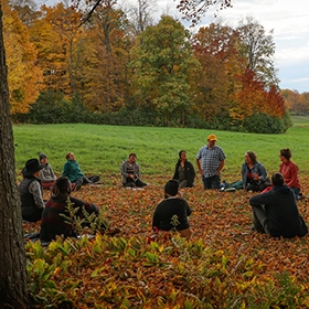 A dozen people sit in a circle on a grassy autumn lawn with fall foliage in distance