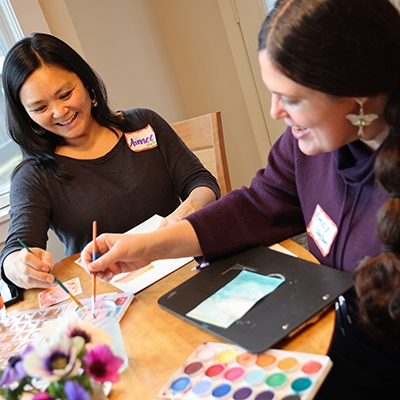 Two women smile while painting with watercolors at a wooden table