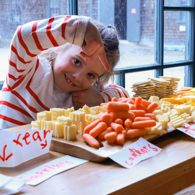 a young girl smiles and points to a large wooden board full of cheese, crackers, carrots, and other cut vegetables