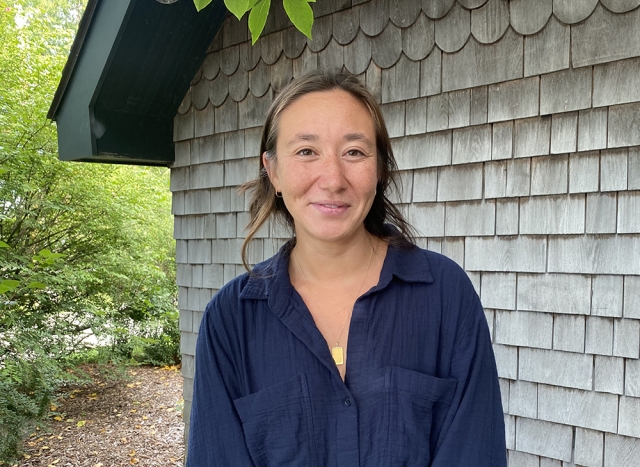 Claire Stoner, in blue shirt, brown hair tied back, and smiling at camera, in front of a shingled building.