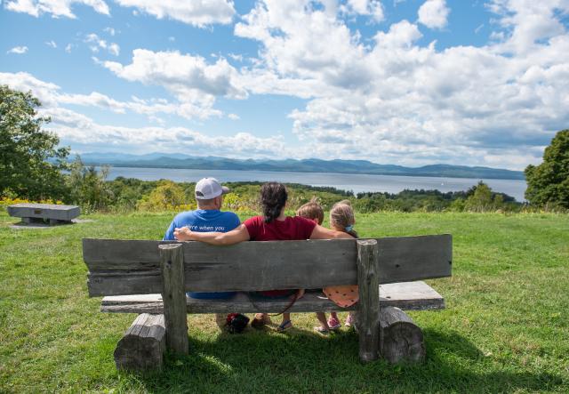 three people on a bench at top of hill overlooking lake champlain and adirondack mountains