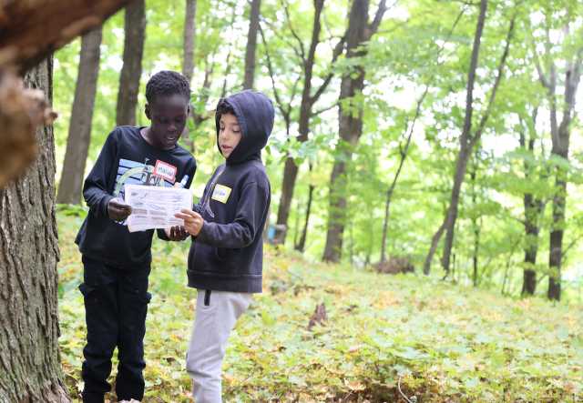 Two young students stand in an autumn forest holding and looking at a piece of paper