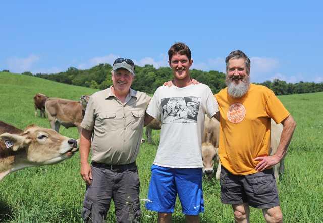 three men stand in green field with cows, the first in baseball cap, the third with gray beard.