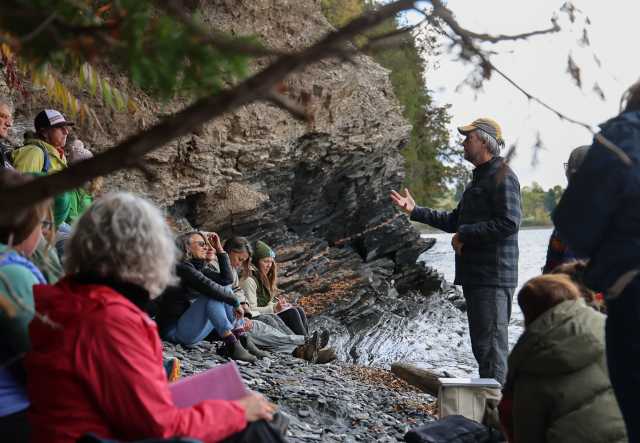 A dozen adults sit and stand along a rocky lakeshore in fall, listening to an instructor speaking to the group
