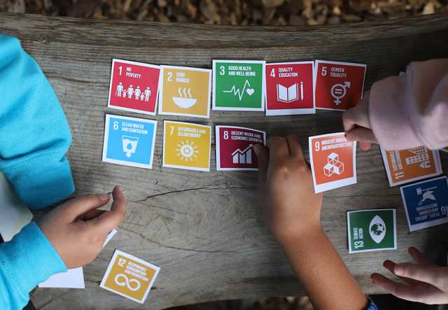 Colorful cards labeled with the UN Sustainable Development Goals are spread out on an outdoor bench as the hands of three young people reach to move the cards