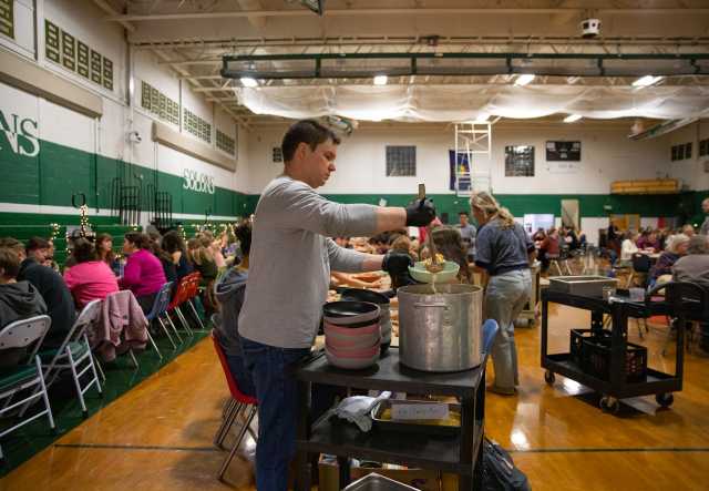 A teacher ladles soup into a bowl. In the background, many students are seated at long tables in a school gymnasium.