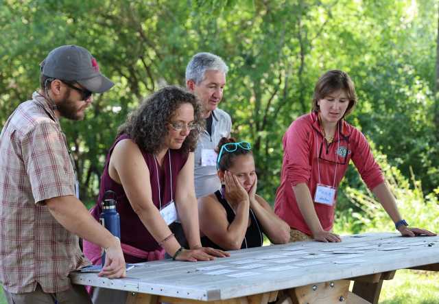 A group of five adults sit and stand around an outdoor table, looking intently at cards spread out on the table surface