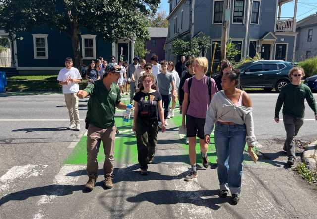 A large group of high school aged students cross a street in discussion with an urban park ranger